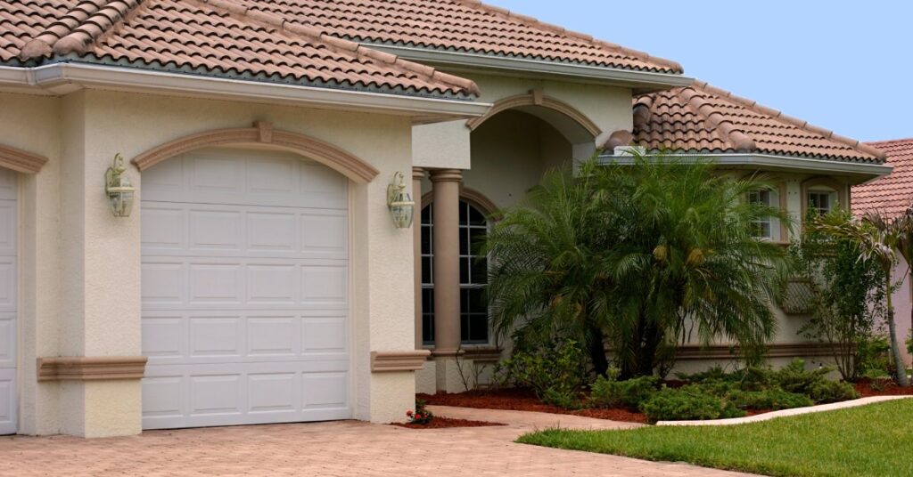 The exterior of a cream-colored house with a reddish-brown roof, a white garage door, and a brick driveway.