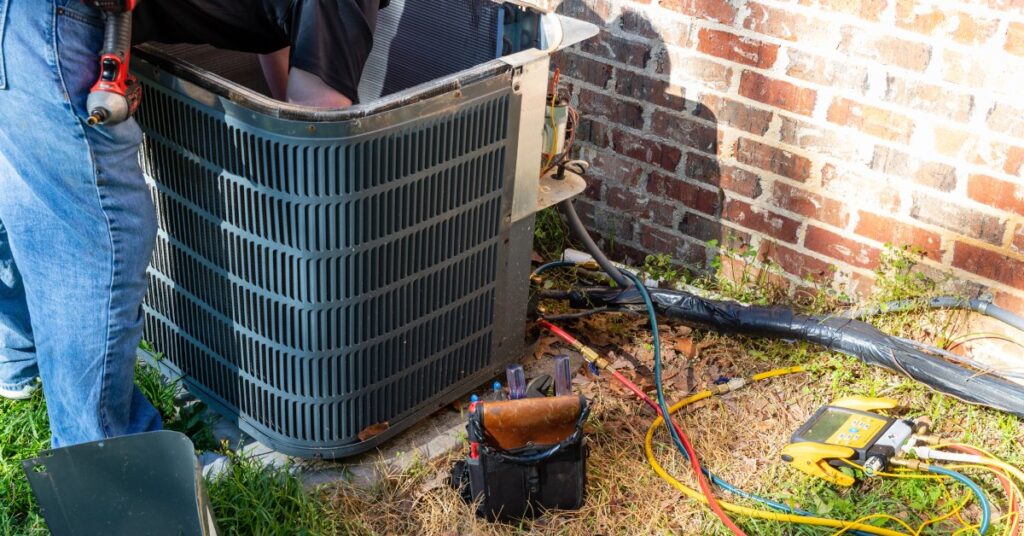 An HVAC tech bending over and reaching inside an outdoor A/C unit. The person carries a power drill at his waist.