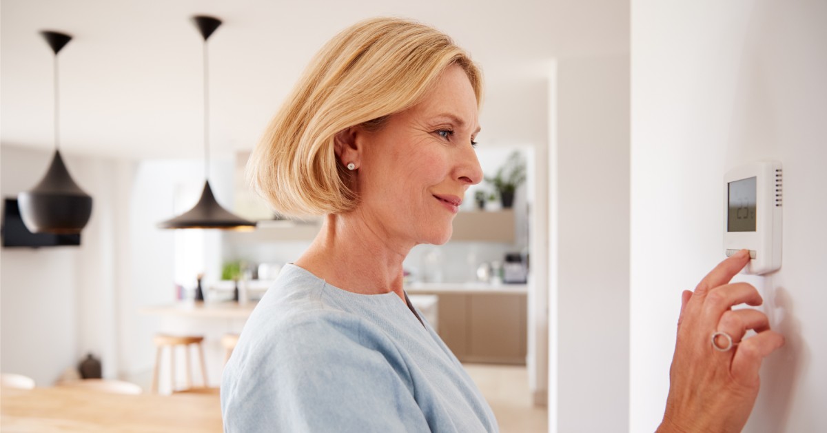 Inside of a home, a woman with short blonde hair stands in front of a thermostat with her finger on the component.