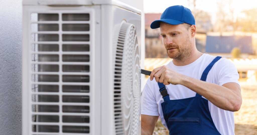 An HVAC tech in a blue hat and industrial apron working on an outdoor AC unit that’s installed on the side of a property.