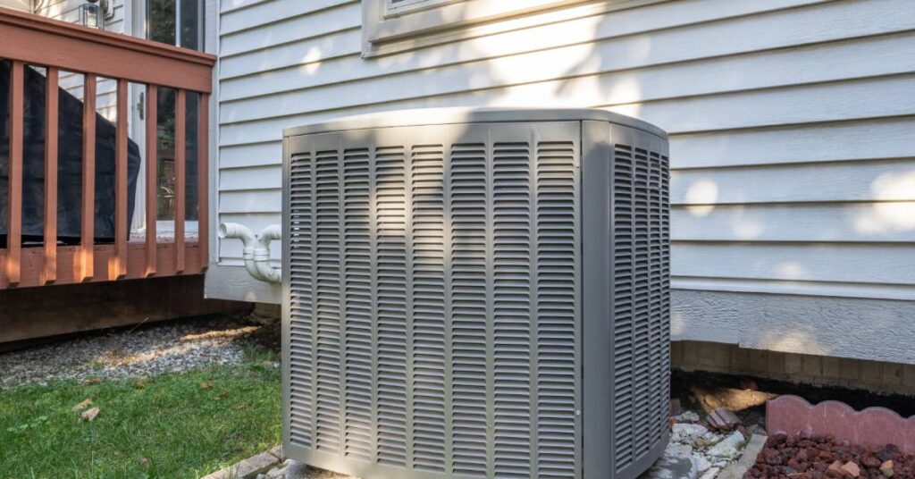 An air conditioning system placed outside, beside the window of a home with white siding; the AC system is in the shade.