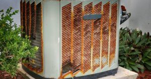 An old and rusted AC unit sits outside of a property beside a bush. The AC unit is on top of a small concrete slab.