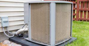 A dirty AC unit that has dust and dirt covering it sits outside of a home with a home that has a wood fence.