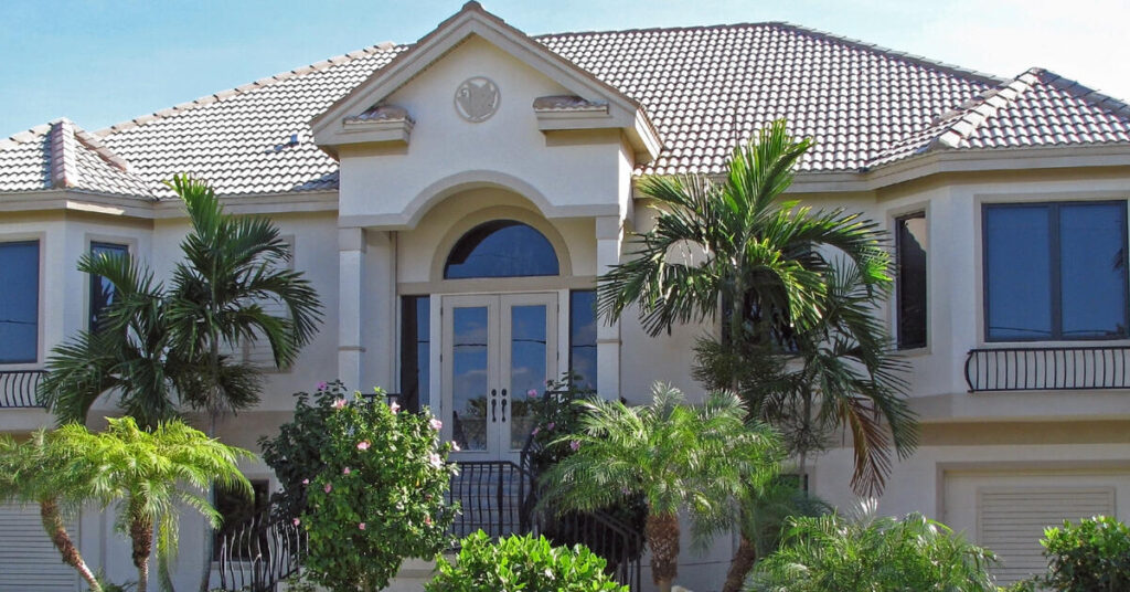 The outside of a beautiful, two-story home that has lush greenery such as palm trees and bushes in the front yard.