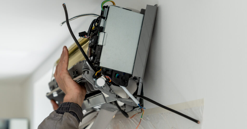 A man looking up at a wall-mounted mini-split unit. His hands are extended up on the unit to hold it in place as he works.