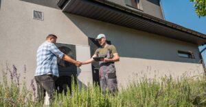 A contractor standing outside of a home beside an HVAC unit shaking the hand of a man in a plaid button-down shirt.