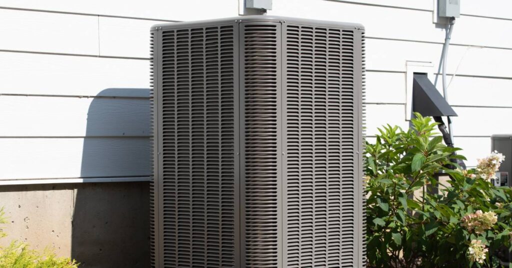 A large, outdoor HVAC unit placed on a concrete slab sits beside a floral bush and a property with white siding.