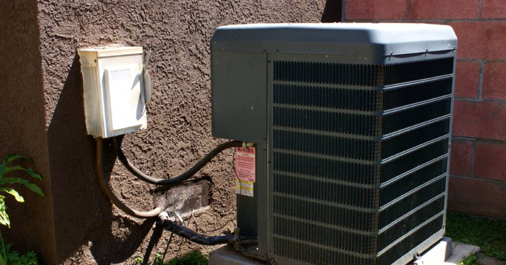 A large air conditioning unit outside of a concrete house. There are hoses on the back of the AC to connect it to the home.