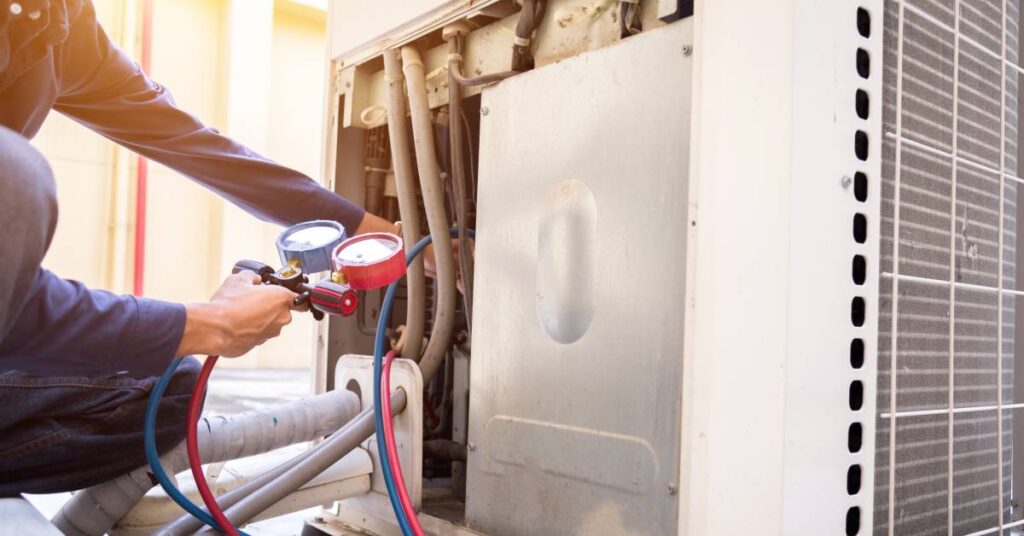 An HVAC technician holds a measurement gauge with blue and red hoses inside of an open AC unit to check the appliance.