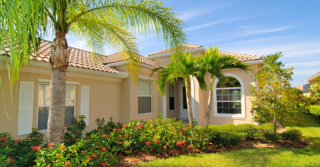 The exterior of a mid-sized, beige-colored home with a brown roof. Several palm trees grow in the front yard.