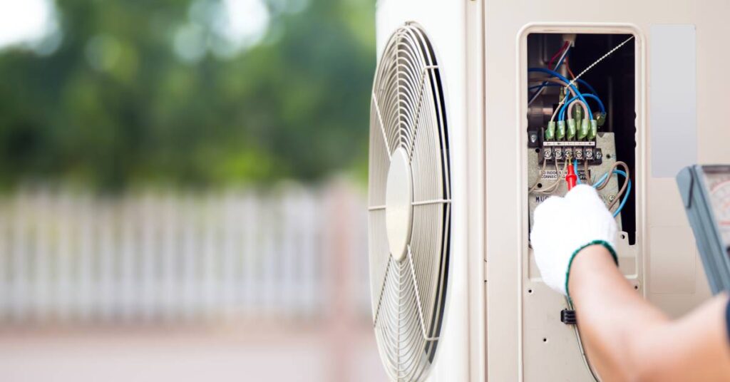 A residential HVAC unit from the side. The access panel is open, exposing the wires. A technician checks the wiring.