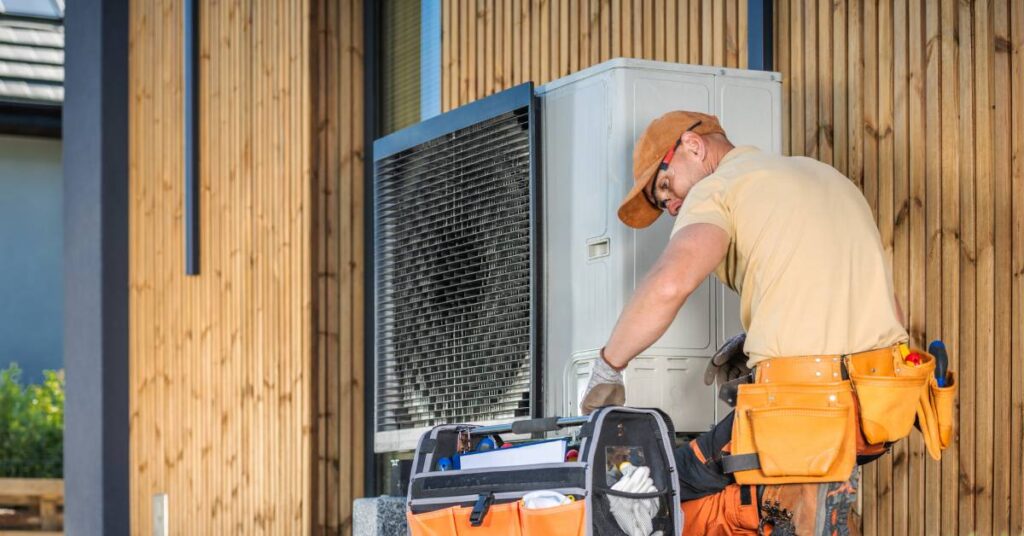 An HVAC worker kneels beside an outdoor, residential AC unit and looks in his tool bag as he services the unit.