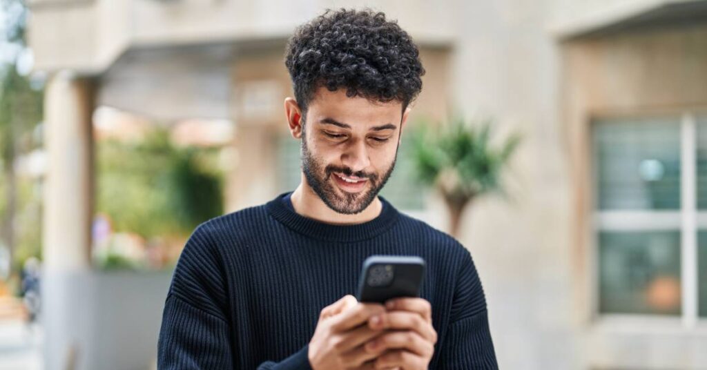 A man with curly hair wearing a black shirt stands outside and holds his cellphone; he smiles as he looks at the screen.