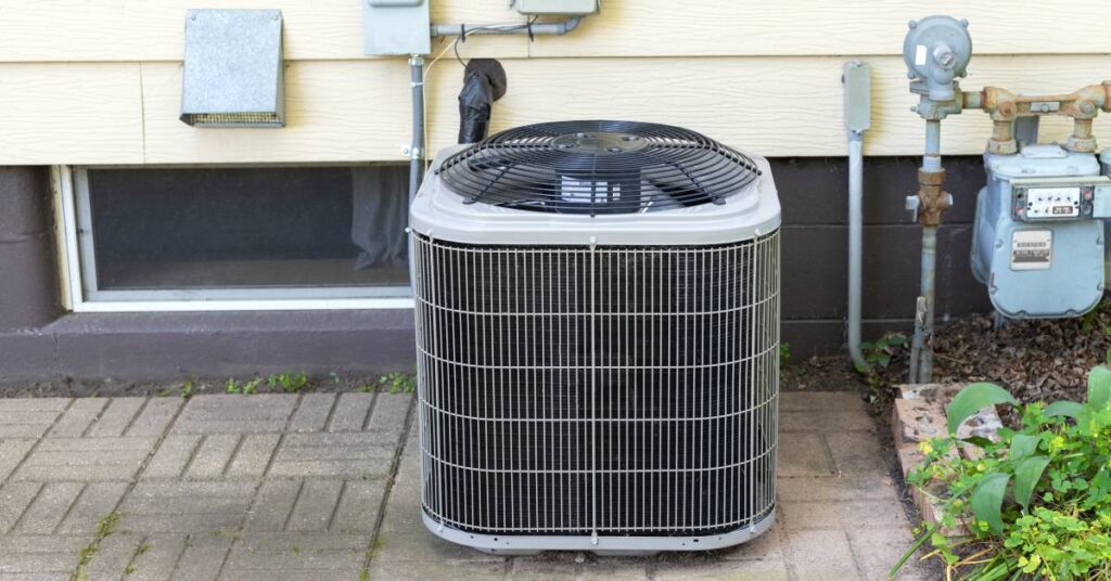 An outdoor, residential-sized AC unit sits on stone pavers beside a home that has cream-colored siding.