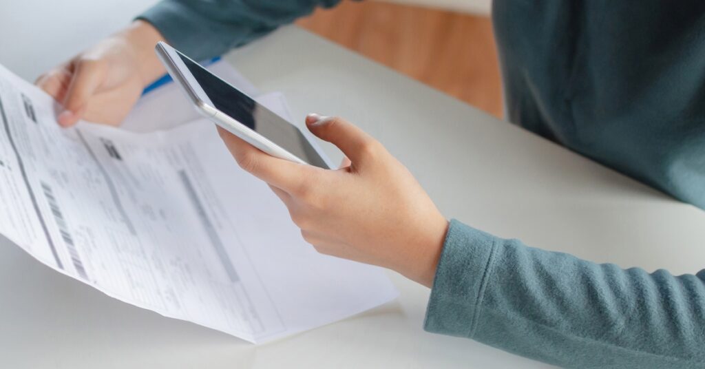 A pair of hands holding sheet of paper with information printed on it; the other hand holds a white smart phone.