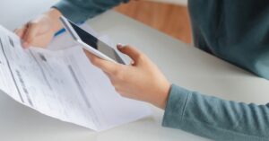 A pair of hands holding sheet of paper with information printed on it; the other hand holds a white smart phone.