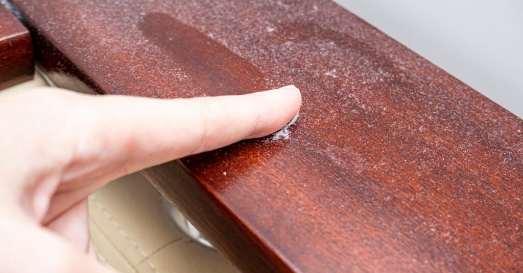 A closeup of someone’s index finger swiping a singly line of dust across a cherry-wood surface inside of a property.