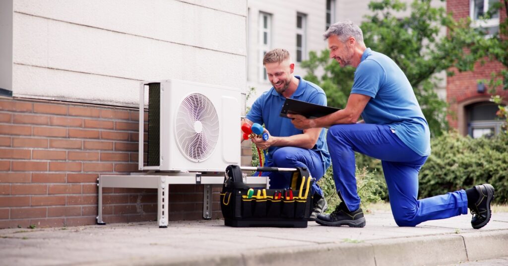 Two men in blue polos and jeans kneel beside an outdoor HVAC unit; there’s a black tool bag beside one of the men.