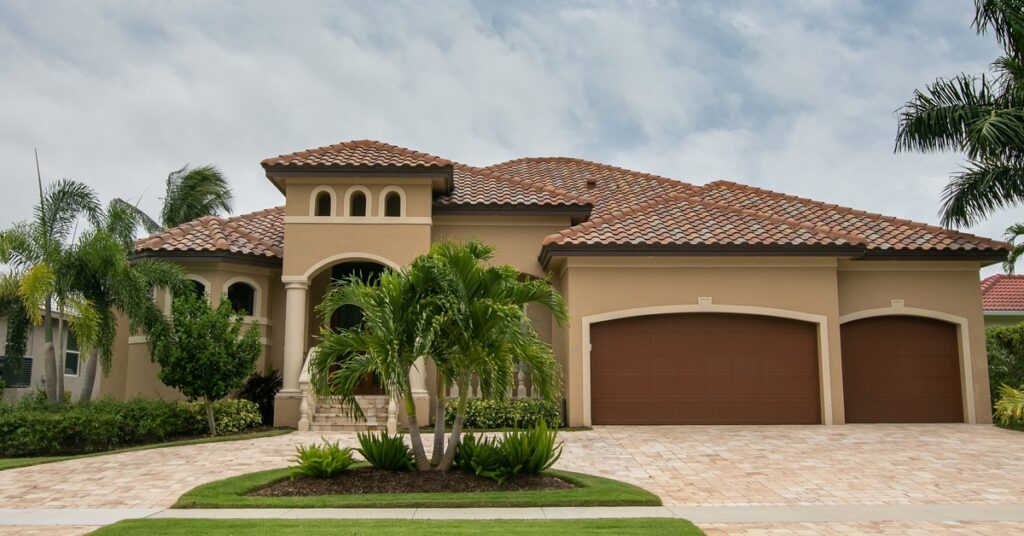 A beautiful, tan home with a clay tile roof and a stone driveway. There are also palm trees and lush grass on the front lawn.