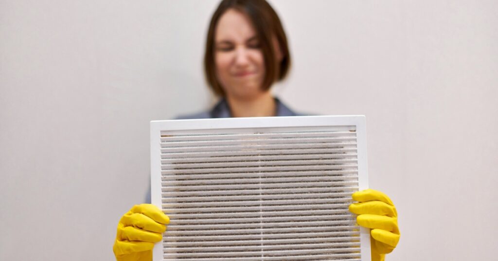 A woman wearing yellow rubber gloves closes her eyes and wrinkles her nose as she holds a dirty HVAC air filter.