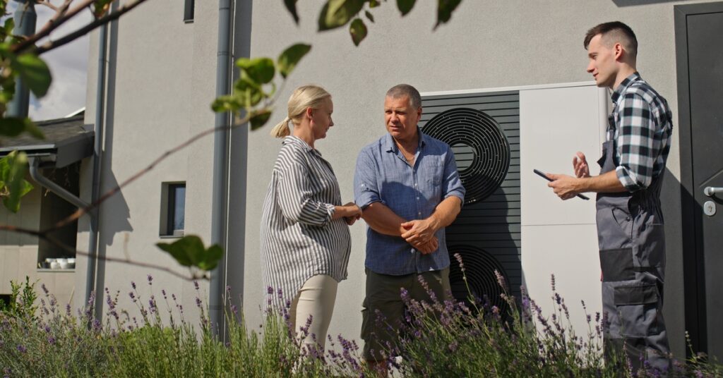 A middle-aged couple and a repair man in overalls stand in front of an outdoor HVAC unit and talk.