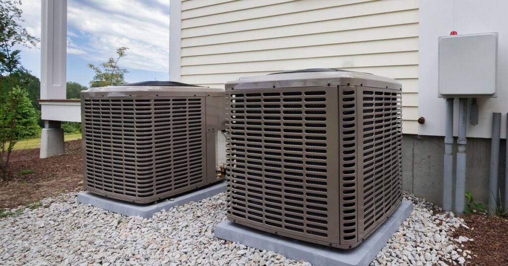 Two HVAC units placed beside one another outside of a white-sided home. The units sit on concrete slaps surrounded by rocks.