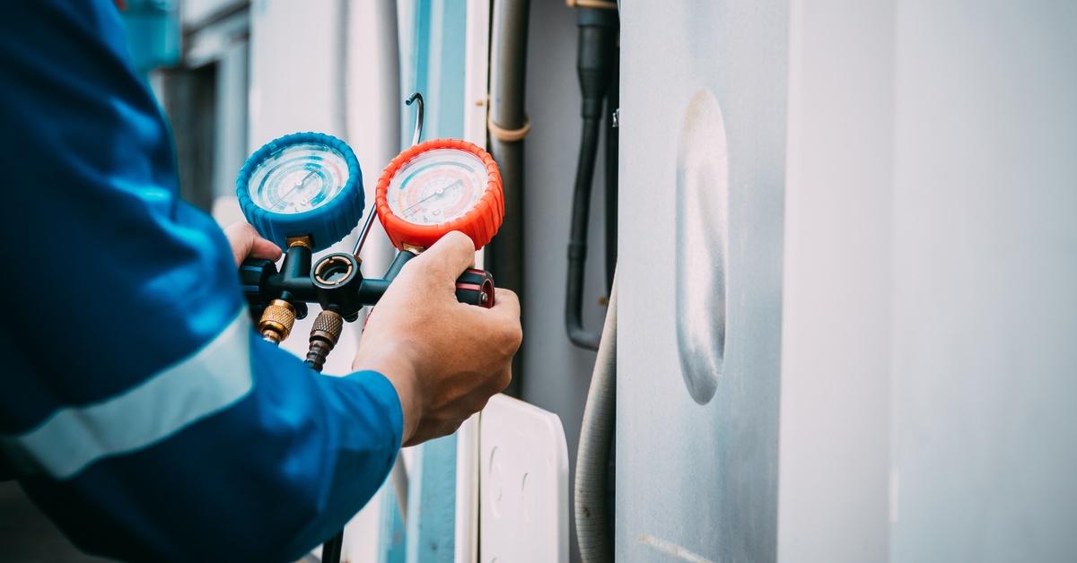 A pair of hands holds a manifold gauge set by an open HVAC unit to check the refrigerant levels of the AC system.