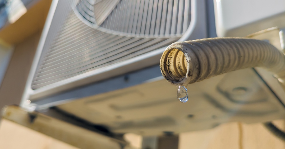 An AC drainpipe with a single drop of water formed at the base; the AC unit is above the pipe and slightly blurred.