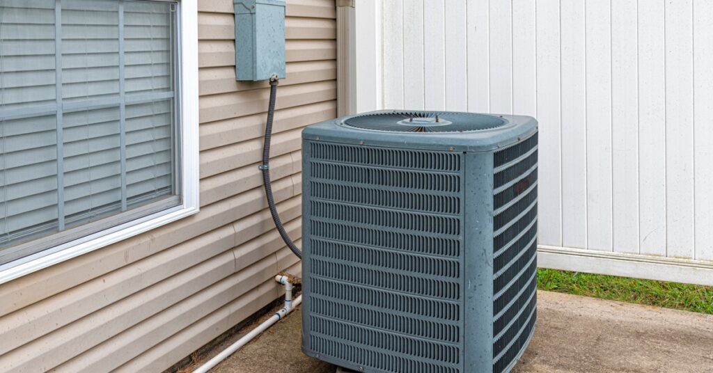 An outdoor HVAC unit sitting on two blocks outside of a home on a large slab of concrete beside a white panel fence.