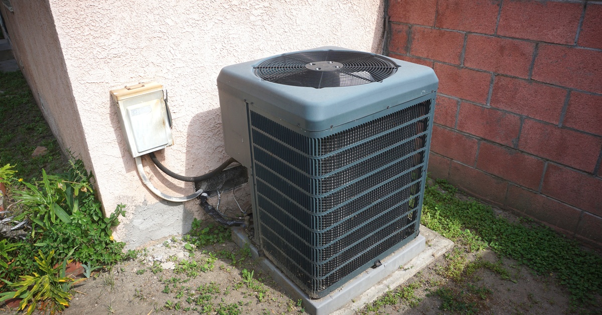 A blue residential outdoor air conditioning unit sits in a corner outside of a building in the sunlight.