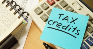 A blue sticky note with the words “tax credits” written on it sits on top of a calculator beside a spreadsheet and ledger.