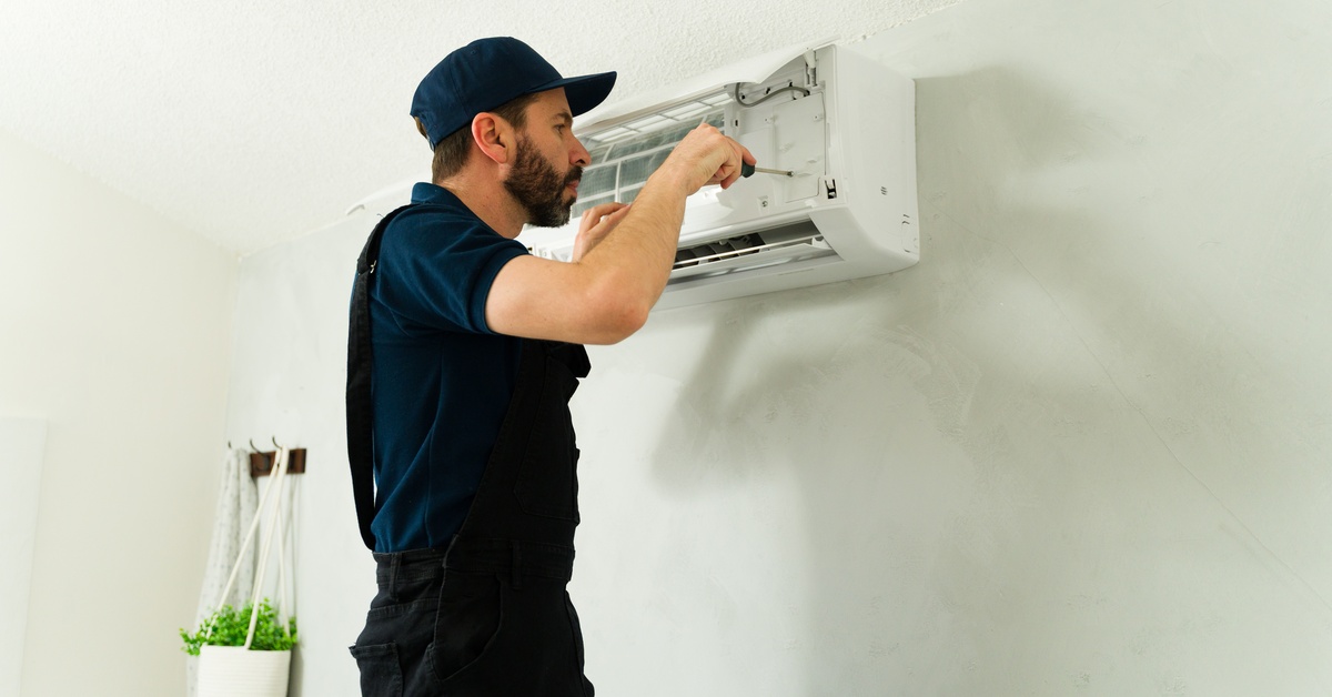 A man in a dark shirt and overalls holds a screwdriver up to an open indoor mini-split for maintenance.