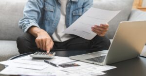 A person in a denim shirt and black pants sitting on a couch by a coffee table with a calculator, laptop, and paperwork.