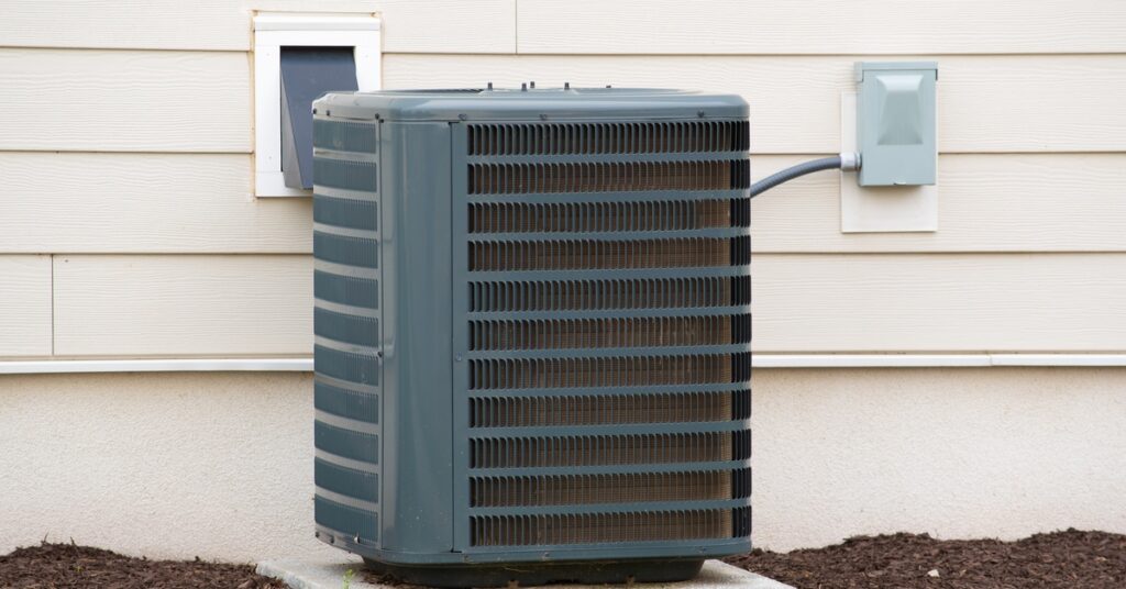 The outdoor half of an AC unit sits on a concrete slab on the mulched perimeter of a white-sided property.