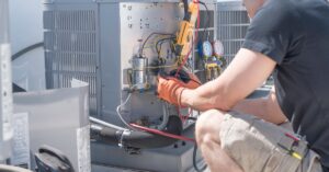 A man in a short-sleeved shirt and shorts kneeling in front of an outdoor HVAC unit with the panel open, exposing wires.