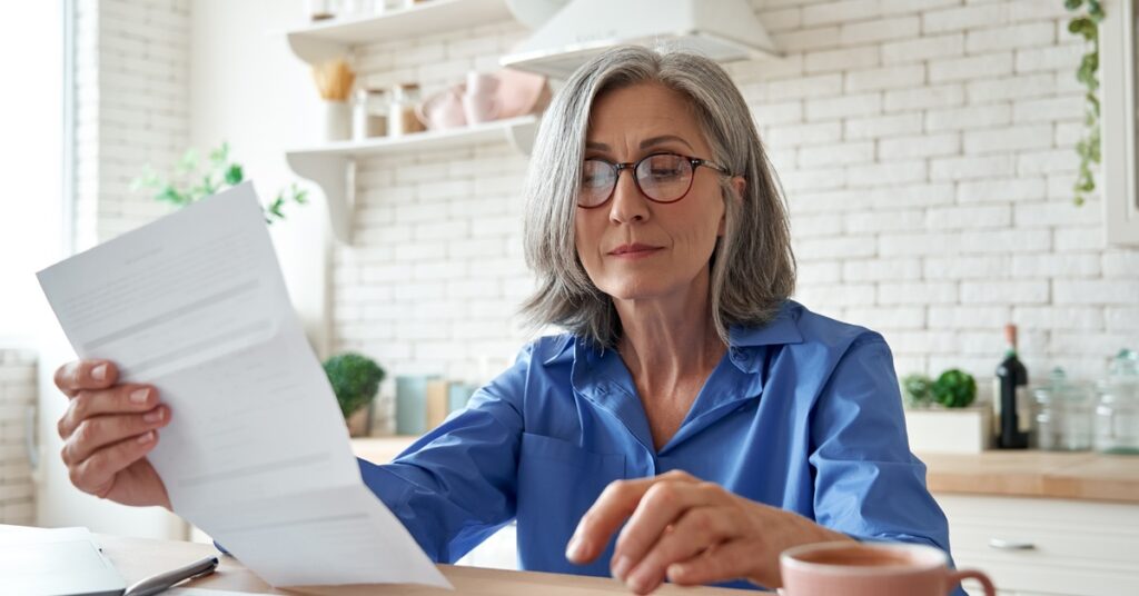 A woman sitting at a table in a white kitchen holding a piece paper in one hand as the other hovers above a calculator.