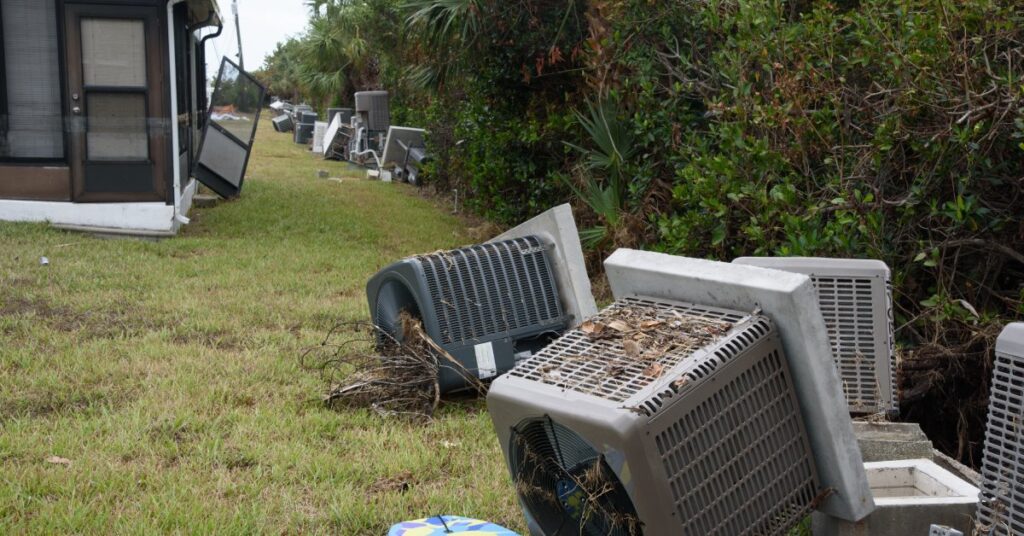 Two outdoor air conditioner units toppled over and lying in the grass. A boogie board is on the ground beside one unit.