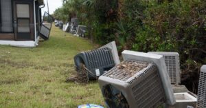 Two outdoor air conditioner units toppled over and lying in the grass. A boogie board is on the ground beside one unit.