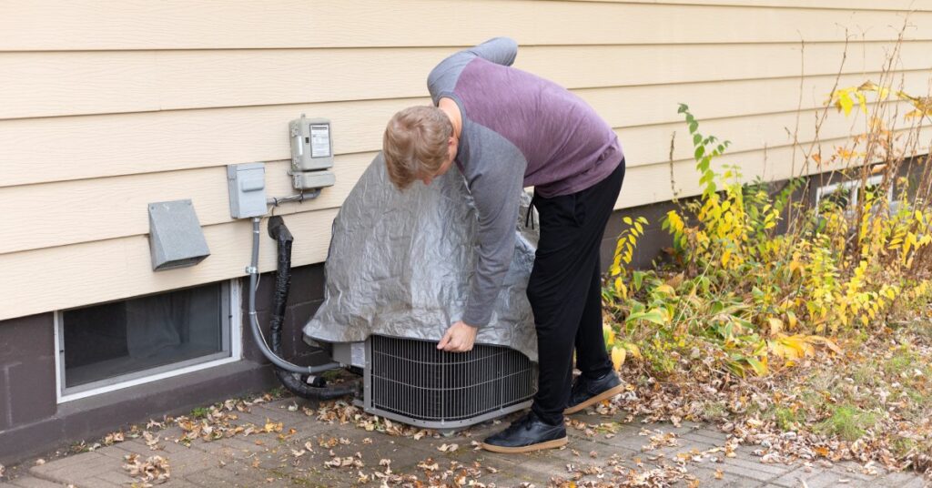 A man in jeans and a long-sleeved shirt placing a gray cover over the outside AC unit that’s beside a home.