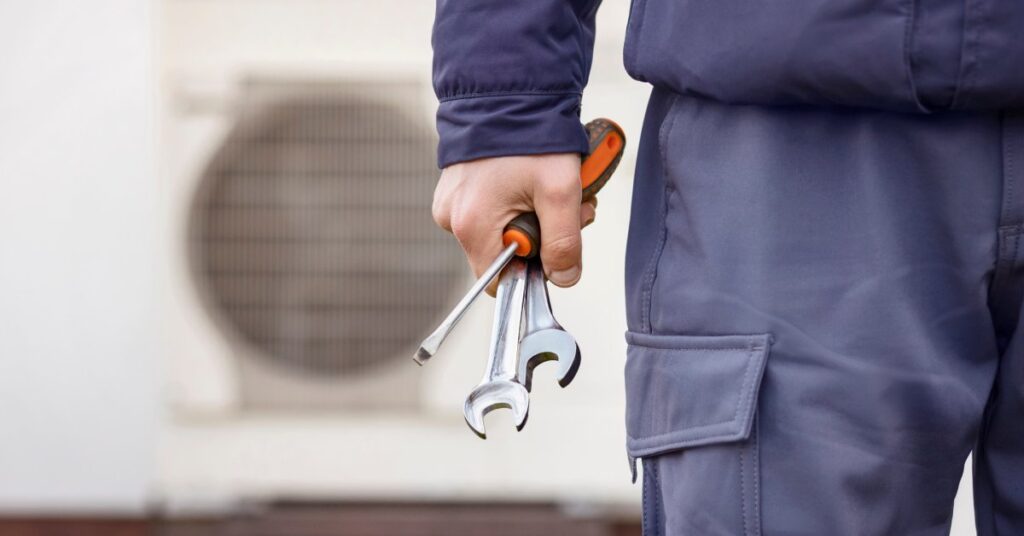 A person in a navy blue jacket and pants holds a screwdriver and two wrenches. An AC unit is blurred in the background.