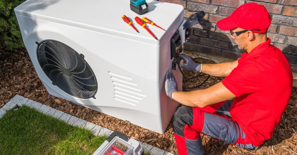 A man kneeling on the ground beside an outdoor AC unit looks at the side of it and has a tool kit beside him.