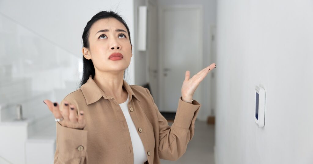 A woman in a tan jacket stands inside by a thermostat as she looks up with both of her hands palm up toward the ceiling.