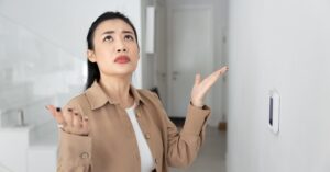 A woman in a tan jacket stands inside by a thermostat as she looks up with both of her hands palm up toward the ceiling.