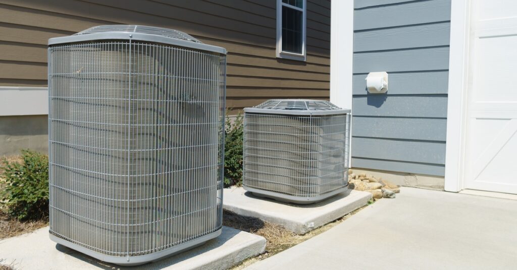 A tall, large AC unit sits on a slab on concrete beside a shorter one; both are outside near residential properties.