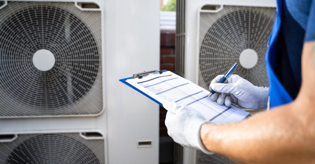 An electrician wearing white gloves stands in front of AC units. He is holding a clipboard with papers.