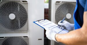 An electrician wearing white gloves stands in front of AC units. He is holding a clipboard with papers.