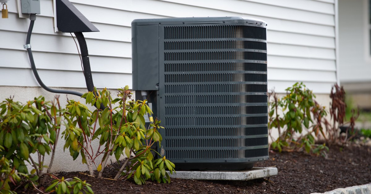 An outdoor air conditioning unit sits on a small concrete slab that’s on top of mulch beside a home with white siding.