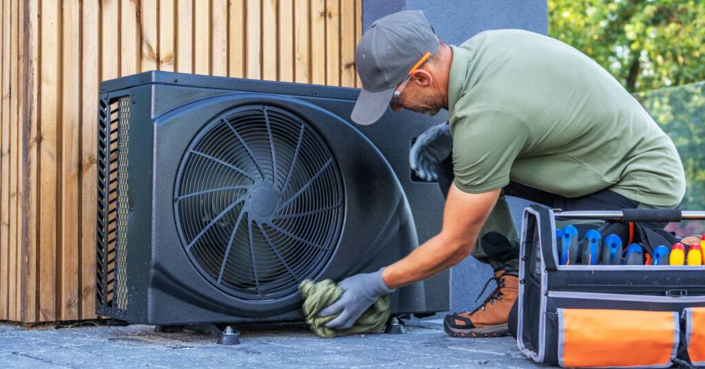 A man holding a rag kneels beside an outdoor HVAC system; there’s a toolbox on the ground beside him.
