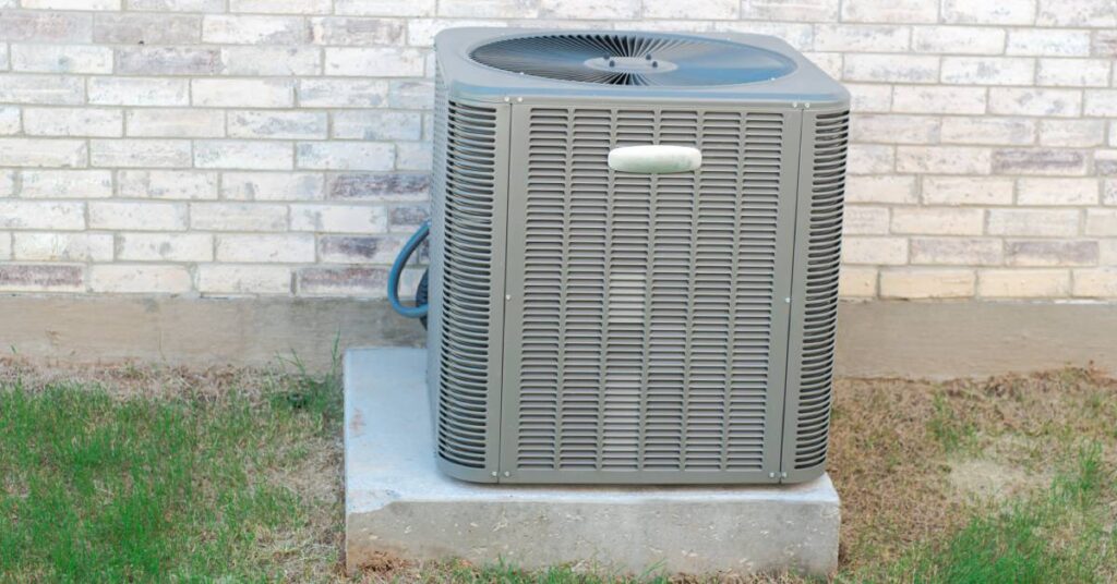 An outdoor HVAC unit on a large cement slab outside of a gray brick building; only patches of grass surround the system.
