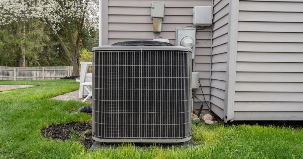 An outdoor air conditioning unit sits in the grass beside a home with gray siding; there’s a tree in the distance.